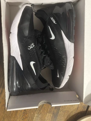 Nike Air Max 270 svartvita sneakers - Nike Air Max 270 sneakers i svart mesh med vita detaljer och stor synlig Air-enhet i hälen. Swoosh-logga på sidan och 270-tryck på plösen. Yttersulan är vit med svart och röd undersida. Skön och sportig design som passar till streetwear.