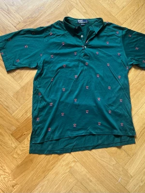 Grön piké med små broderade mönster - Grön pikétröja från Polo Ralph Lauren med korta ärmar och små broderade emblem i rött och vitt över hela tröjan. Klassisk krage och knappar framtill. Tillverkad i mjuk bomull för en bekväm känsla.