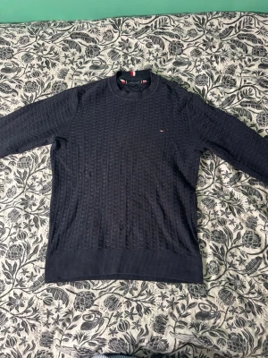 Mörkblå stickad tröja Tommy Hilfiger - Snygg mörkblå stickad tröja från Tommy Hilfiger med diskret logga på bröstet. Tröjan har rund halsringning, ribbade muddar och ett strukturerat stickmönster. Perfekt för dig som gillar klassisk och stilren design.