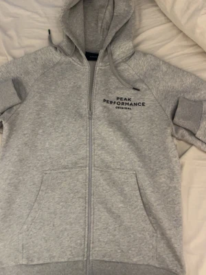 Grå hoodie Peak Performance - Snygg ljusgrå hoodie från Peak Performance med dragkedja framtill och två stora fickor. Klassisk logga på bröstet och justerbar huva med snören. Tillverkad i mjukt sweatshirtmaterial, perfekt för chill dagar.