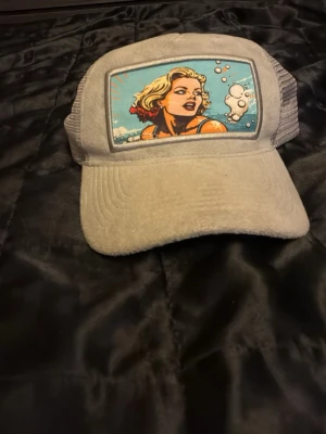 Grå truckerkeps med pop art-tryck - Säljer en grå truckerkeps med mesh på sidorna och ett färgstarkt pop art-tryck framtill som föreställer en blond person i vatten med pratbubbla. Kepsen har böjd skärm och justerbar snapback. Perfekt för dig som gillar streetwear och unika accessoarer.