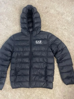 Svart dunjacka från EA7 Emporio Armani - Säljer en svart dunjacka från EA7 Emporio Armani med huva och dragkedja framtill. Jackan har ett stilrent, quiltat mönster och logotyp på bröstet. Perfekt för kalla vinterdagar och har två sidofickor med dragkedja.