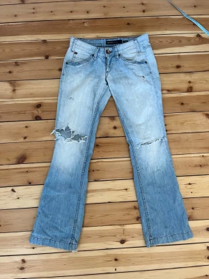 Ljusblå bootcut jeans från Miss Sixty hål revor - Säljer ett par ljusblå bootcut jeans från Miss Sixty med slitningar och hål på knäna. Jeansen har klassisk femficksmodell, låg midja och detaljerade bakfickor. Tillverkade i bomull med en cool vintagekänsla.