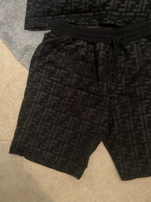 Svarta Fendi shorts och t-shirt med logomönster - Snygga svarta shorts från Fendi med det klassiska FF-logomönstret över hela plagget. Shortsen har snörning i midjan och är tillverkade i ett mjukt material som känns bekvämt. Perfekta för dig som vill ha en exklusiv och trendig look.