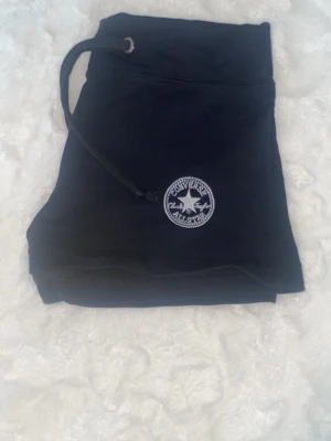 Svarta Converse shorts med snörning - Svarta shorts från Converse med klassisk logga på vänster sida. Modellen har hög midja och snörning i midjan för justerbar passform. Perfekta för sommardagar eller chill hemma. Mjukt material som känns skönt mot huden.