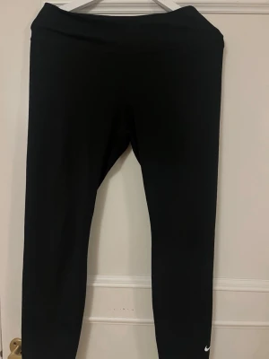 Svarta leggings från Nike - Säljer ett par svarta leggings från Nike med klassisk vit logga vid vristen. De har hög midja och är tillverkade i ett stretchigt material som sitter tight och bekvämt. Perfekta för träning eller chill dagar. Passar dig som gillar sportig och enkel stil.