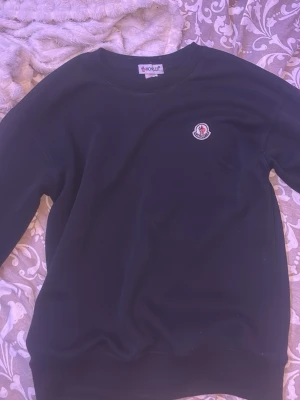 Svart sweatshirt från Moncler - Svart sweatshirt från Moncler med rund halsringning och Moncler-logga på bröstet. På ena ärmen finns en stor vit Moncler M-print tillsammans med logotypen. Perfekt för en clean och stilren look. Sliten vid handleden men de går att laga 