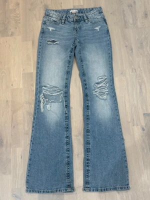 Blå slitna bootcut jeans - Säljer ett par blå bootcut jeans med slitningar och hål på både fram- och baksida. Jeansen har klassisk femficksmodell, låg midja och råa kanter vid bensluten. Perfekta för en avslappnad och trendig look. Storlek xs tall, innerbenslängden är ca 85cm