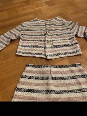 Randigt set med kjol och jacka - Säljer ett snyggt tvådelat set med kjol och jacka i beige, svart och ljusrosa ränder. Jackan har långa ärmar, rund halsringning, dekorativa fickor och knappar framtill. Kjolen är rak och kort. Materialet är vävt och ger en trendig, lite retro känsla.