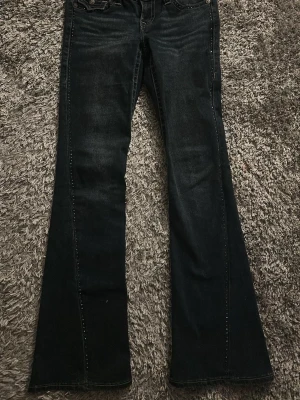 True Religion Joey Low Rise Flare Jeans - Säljer ett par mörkblå True Religion Joey jeans med low rise och flare passform. Klassiska kontrastsömmar och kristaller och snygg tvätt. Perfekta för dig som gillar retrovibbar och vill ha ett par jeans som sticker ut lite extra.