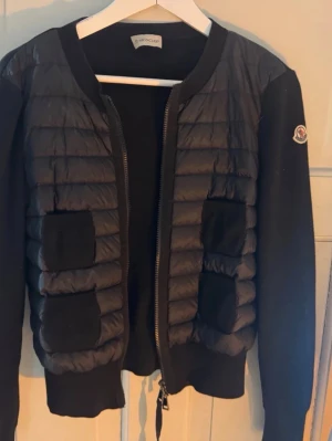 Svart cardiganjacka från Moncler - Snygg svart cardiganjacka från Moncler med quiltad framsida och mjuka stickade ärmar. Jackan har dragkedja framtill, två fickor och Moncler-logga på ärmen. Perfekt för lager-på-lager och en sportig, trendig look.