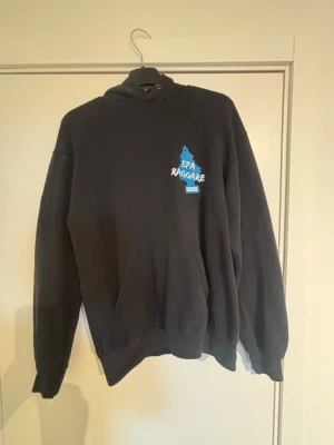 Svart hoodie EPA Raggare tryck - Svart hoodie med stor blå EPA Raggare-tryck på ryggen och mindre tryck på bröstet. Klassisk känguruficka framtill och huva med dragsko. Tillverkad i mjukt bomullsmaterial, perfekt för dig som gillar streetwear och vill sticka ut.