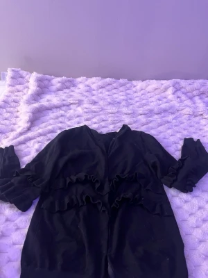 Svart volangkofta med långa ärmar - Snygg svart kofta med volangdetaljer längs bröstet och ärmarna. Koftan har en croppad passform och långa ärmar, perfekt för att lyfta en enkel outfit. Tillverkad i mjukt material som känns skönt mot huden.