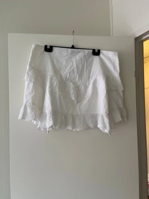 Vit volangkjol med spetsdetaljer - Supersöt vit kortkjol med volanger och broderade spetsdetaljer. Kjolen har en luftig känsla och passar perfekt till varma dagar. Tillverkad i bomull för en skön och lätt känsla. Perfekt att matcha med en enkel topp eller stickad tröja.