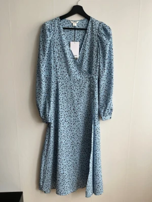 Blommig blå omlottklänning Monki - Säljer en ljusblå omlottklänning från Monki med svart blommigt mönster. Klänningen har långa puffärmar, v-ringning och knyt i midjan. Perfekt för vår och sommar, tillverkad i ett mjukt och lätt material. Sitter tajt runt midjan men lös i armer och kjolen. Aldrig använd! 