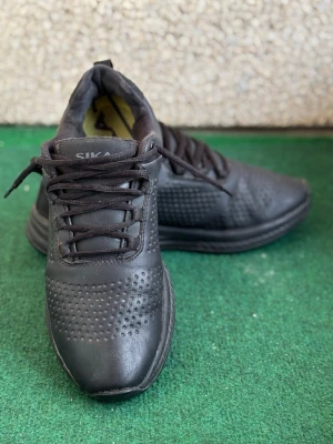 Svarta sneakers från Sika - Svarta sneakers från Sika med perforerade detaljer på ovandelen och klassisk snörning. Skorna har en robust svart sula med mönster och gul detalj undertill. Ovandelen är i syntetmaterial och designen är sportig och stilren.