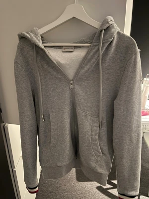 Grå zip hoodie från Moncler - Snygg grå hoodie från Moncler med dragkedja framtill, två fickor och justerbar huva med snören. Ribbstickade muddar med röd och marinblå detalj på ärmsluten. Perfekt för en chill och avslappnad stil.