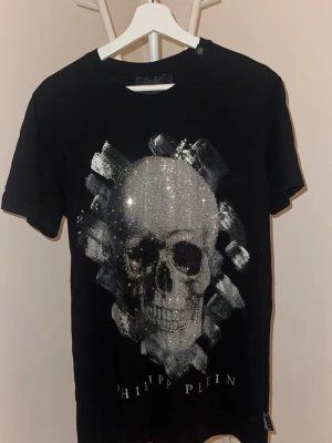 Svart t-shirt från Philipp Plein - Snygg svart t-shirt från Philipp Plein med ett coolt tryck i silver och vitt framtill. T-shirten har rund halsringning och korta ärmar. Tillverkad i mjuk bomull som känns skön mot huden. Perfekt för dig som gillar streetwear och vill sticka ut.
