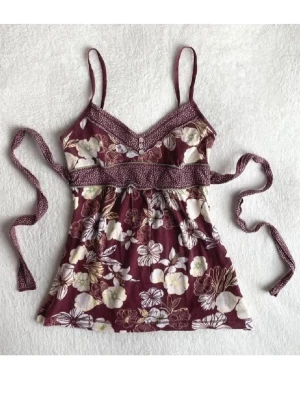 Blommig linnetopp från Hollister - Supersöt linnetopp från Hollister i vinrött med blommigt mönster i vitt och beige. Toppen har smala axelband, v-ringning och knytband i midjan för extra snygg passform. Perfekt för varma dagar och sommarvibbar! OBS långa band! Behöver förmodligen sys