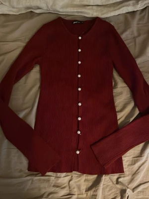 Vinröd kofta från Gina Tricot - Säljer en vinröd ribbad kofta från Gina Tricot med långa ärmar och pärlknappar framtill. Koftan har en smal passform och rund halsringning, perfekt att styla till olika outfits.