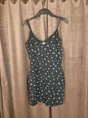 Svart blommig kortklänning med spets - Svart kortklänning från H&M med vita små blommor och tunna axelband. Dekorerad med spetskant vid urringningen och en liten rosett framtill. Mjuk och stretchig känsla, perfekt för sommaren.