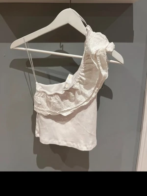 Vit oneshoulder topp med volang Gina Tricot - Superfin vit oneshoulder topp från Gina Tricot med broderad volangdetalj. Tunn axelrem på ena sidan och bred volang på andra ger en unik look. Perfekt till sommaren och lätt att matcha med jeans eller kjol.