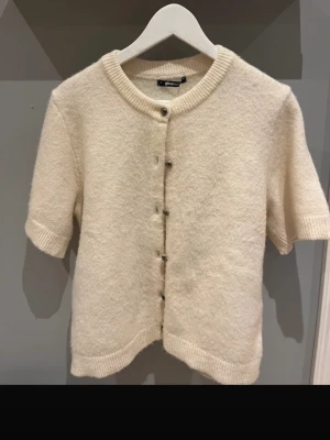 Krämvit kofta från Gina Tricot - Kortärmad krämvit kofta från Gina Tricot med rund halsringning och ribbade kanter. 🧨STORLEK XL MEN PASSAR S🧨Koftan har knappar framtill och är gjord i ett mjukt, fluffigt material som känns riktigt cozy. Perfekt att slänga över en topp för en chill look.