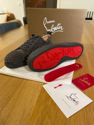 Christian Louboutin svarta spikes sneakers - Svarta sneakers från Christian Louboutin med klassiska röda sulor och svarta nitar över hela tån. Skorna är låga och har en lyxig känsla med sin matta mocka och unika design. Perfekta för dig som vill sticka ut med exklusiva detaljer och edgy look.