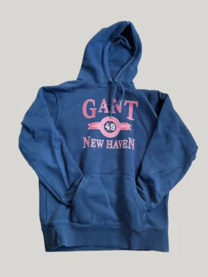 Gant new heaven hoddie - Snygg marinblå hoodie från Gant med tryck i rött och vitt på bröstet där det står 'GANT 49 NEW HAVEN'. Klassisk känguruficka framtill och justerbar huva med dragsko. Perfekt för en avslappnad och sportig stil.
