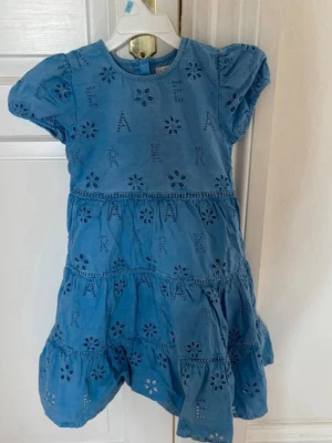 Blå broderad klänning med puffärm - Söt blå klänning med broderade blommor och bokstäver, korta puffärmar och rund hals. Klänningen har volangkjol och är tillverkad i ett lätt bomullstyg som känns luftigt och somrigt. Perfekt för dig som gillar en lekfull och avslappnad stil.