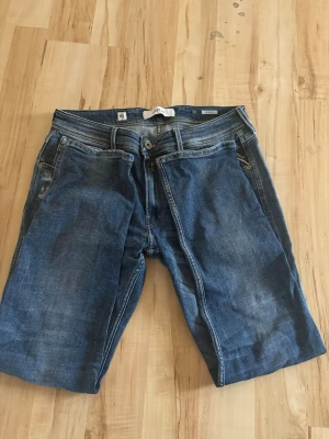 Blå slitna replay jeans med raka ben - Säljer ett par blå jeans med raka ben och slitningar på framsidan. Jeansen har klassisk femficksdesign, knappgylf och är tillverkade i denim. Perfekta för en avslappnad och trendig look.