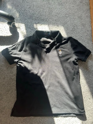 Svart pikétröja från Polo Ralph Lauren - Klassisk svart pikétröja från Polo Ralph Lauren med broderad björnlogga på bröstet. Tröjan har krage, korta ärmar och knappar vid halsen. Tillverkad i mjukt bomullsmaterial som är skönt mot huden. 