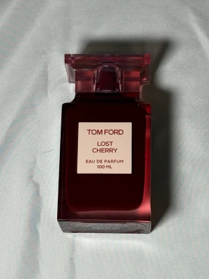 Tom Ford Lost Cherry EdP 100 ml - Tom Ford Lost Cherry Eau de Parfum i en lyxig, mörkröd glasflaska med rosa transparent kork. Flaskan rymmer 100 ml och har en elegant, fyrkantig form med en tydlig etikett framtill. Doften är känd för sina söta och fruktiga toner.