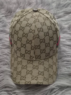 Beige keps från Gucci med GG-mönster - Säljer en beige keps från Gucci med det klassiska GG-monogrammet över hela kepsen. På sidan finns en grön och röd rand som ger extra lyxig känsla. Kepsen är tillverkad i canvasmaterial och har böjd skärm. Perfekt för dig som gillar exklusiva accessoarer.