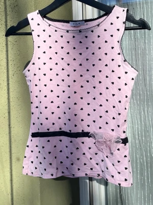 Rosa linne med hjärtan från Vila - Supersött rosa linne från Vila med svarta små hjärtan över hela tyget. Dekorativ spetskant vid halsringningen och ett svart band med rosett och tyllvolang i midjan. Perfekt för dig som gillar gulliga detaljer och en lekfull stil.