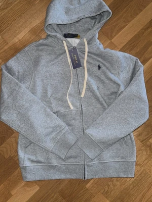 Grå hoodie från Polo Ralph Lauren - Snygg grå hoodie från Polo Ralph Lauren med dragkedja, vit dragsko och klassisk logga broderad på bröstet. Tillverkad i mjuk bomullsblandning och har känguruficka framtill. Perfekt för en chill och avslappnad stil.