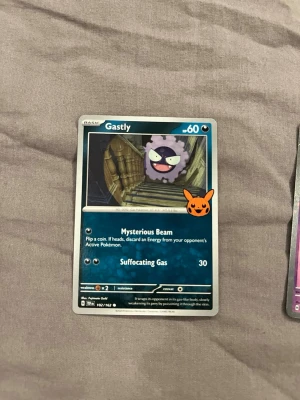 Pokémonkort Gastly med Pikachu-stämpel - Samlarobjekt! Ett Pokémonkort med Gastly-motiv och en orange Pikachu-stämpel på framsidan. Kortet har blå bakgrund och visar Gastly i en trappa. Text och attacker på engelska. Perfekt för dig som gillar Pokémonkort och samlar på unika kort.