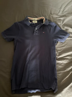 Mörkblå pikétröja Tommy Hilfiger - Klassisk mörkblå pikétröja från Tommy Hilfiger med korta ärmar och liten logga på bröstet. Tröjan har krage och knappar framtill. Perfekt till jeans eller shorts för en avslappnad stil.