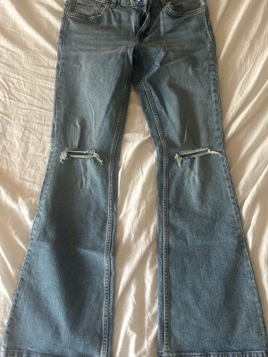Blå bootcut jeans från JC 157 - Säljer ett par blå bootcut jeans från JC 157 med slitningar över knäna. Jeansen har låg midja och klassisk femficksdesign. Perfekta för dig som gillar en avslappnad och trendig look.