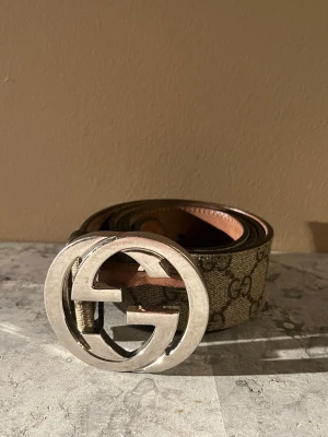 Gucci GG-bälte med silverfärgat spänne - Snyggt Gucci-bälte i beige och brunt med klassiskt GG-mönster och stort, rundat spänne i silverfärg. Bältet är gjort i skinn och har tydliga Gucci-detaljer på insidan. Perfekt accessoar för att lyfta vilken outfit som helst.