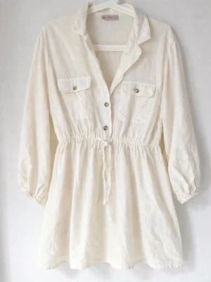 Ljus beige/cremefärgad strandklänning M - Ljus beige/cremefärgad strandklänning / beach cover-up i lätt och luftig modell. Skjortstil med knappar framtill, bröstfickor och markerad midja med resår. Perfekt som påsläng över bikini på stranden eller vid poolen. Kort modell – passar bäst för dig som är ca 160 cm eller kortare. Mycket fint skick.  Storlek: M (ca) Pris: 150 kr