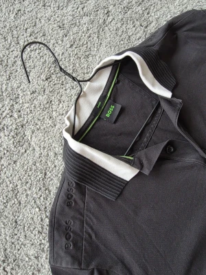 Svart polotopp från BOSS med vit krage - Säljer en svart polotopp från BOSS med vit kontrastkrage och diskret logga på bröstet och axeln. Toppens material är mjuk bomull och den har knappar framtill. Perfekt för dig som gillar stilrena detaljer och klassisk design.