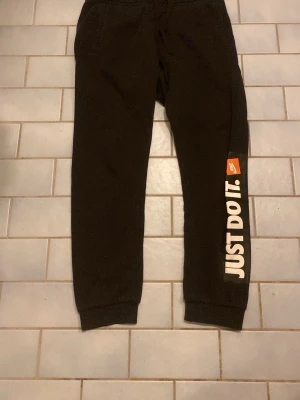 Svarta Nike mjukisbyxor Just Do It - Svarta mjukisbyxor från Nike med tryck 'Just Do It.' längs ena benet och klassisk Nike-logga. Byxorna har resår i midjan och muddar vid benslut, perfekt för chill eller träning. Materialet är mjukt och bekvämt, troligen bomull eller polyester.