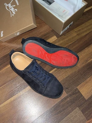 Mörkblå Louboutin sneakers i mocka - Snygga mörkblåa sneakers från Christian Louboutin i mjuk mocka med klassisk röd sula och diskret broderad logga på hälen. Skorna har mörkblåa skosnören och en låg silhuett som ger en clean och exklusiv look. Dessa är för små nu storlek 42 men passar förmodligen bäst för 41