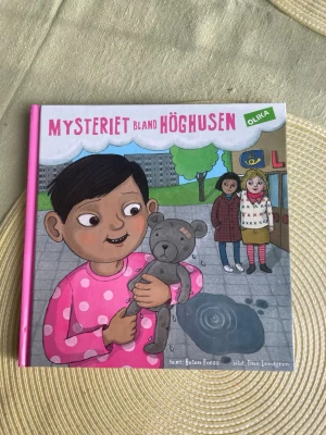 Mysteriet bland höghusen - En färgglad barnbok med hårda pärmar, skriven av Helen Forss och illustrerad av Tina Landgren. Boken handlar om Vilhelm och mysteriet med nallen, och har illustrationer med barn i stadsmiljö. Perfekt för unga läsare som gillar spännande berättelser.