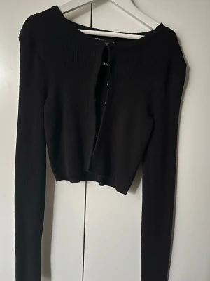 Svart ribbad cardigan - Trendig svart ribbstickad cardigan från FB Sister med croppad passform och långa ärmar. Tröjan har två dekorativa hakar framtill istället för knappar och är tillverkad i mjukt stickat material. Perfekt att styla över ett linne eller t-shirt. Endast använd 1 gång, därav bra skick