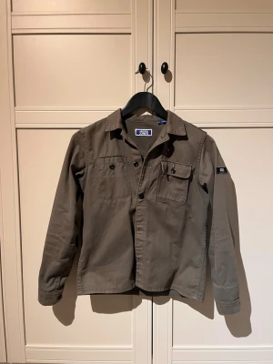 Olivgrön overshirt från Jack & Jones - Snygg oliv overshirt från Jack & Jones med klassisk krage, knappstängning och bröstficka med lock. Tillverkad i ett slitstarkt bomullstyg och har en diskret logotyp på ärmen. Perfekt lager-på-lager-plagg för en avslappnad stil.           Vid snabb o smidig affär  affär kan pris diskuteras 