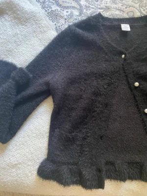 Svart kofta med pärlknappar - Säljer en svart kofta från Lindex med v-ringning och dekorativa pärlknappar framtill. Koftan har långa ärmar och en mjuk textur. 