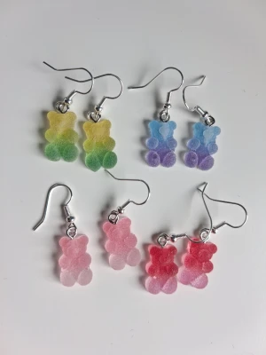 Söta örhängen med nallebjörnar 4st - Handgjorda örhängen formade som små sockriga nallebjörnar i olika färger: gulgrön, blå-lila, ljusrosa och rödrosa. Mått på björnarna är ca 1x1,6cm. Öronkroken är nickelsäker. Blir en söt och gullig accessoar till outfiten 