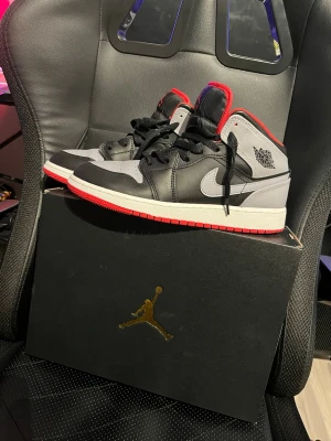 Air Jordan 1 Mid Svart/Röd/Grå - Snygga Air Jordan 1 Mid sneakers i svart, grått och rött med klassisk Jumpman-logga på plösen och vingar på sidan. Skorna har svart läder, gråa detaljer och röd kant runt kragen samt yttersula.